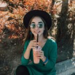 Young woman in sunglasses enjoying a cool beverage outdoors amidst autumn foliage.